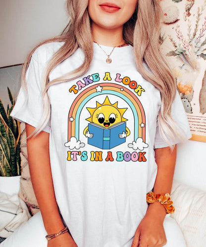 A bright and joyful graphic with a cute sun character reading a book under a vibrant rainbow The phrase take a look it&#39;s in a book is written in bold letters creating a happy and educational message