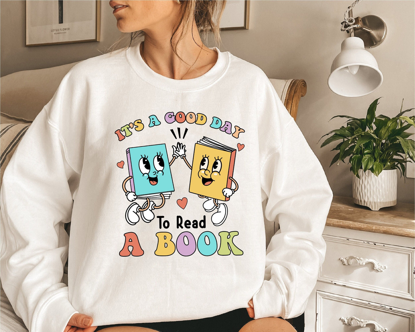 the image shows two books with animated expressions and cartoonish appendages, celebrating the act of reading with a playful high-five, while the phrase IT&#39;S A GOOD DAY TO READ A BOOK is splayed out in cheerful, colorful letters.