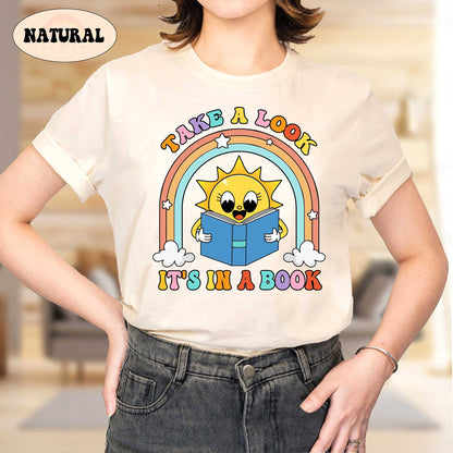 A cheerful design featuring a smiling sun holding an open book with a colorful rainbow arching above The text take a look it&#39;s in a book is displayed in playful fonts adding a fun and inviting vibe