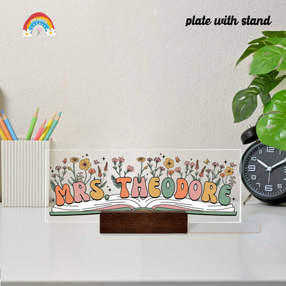 A beautifully crafted nameplate with the name Mrs. Theodore is illustrated with a floral scene, which blooms from a central book, representing the teacher&#39;s role in nurturing student growth.
