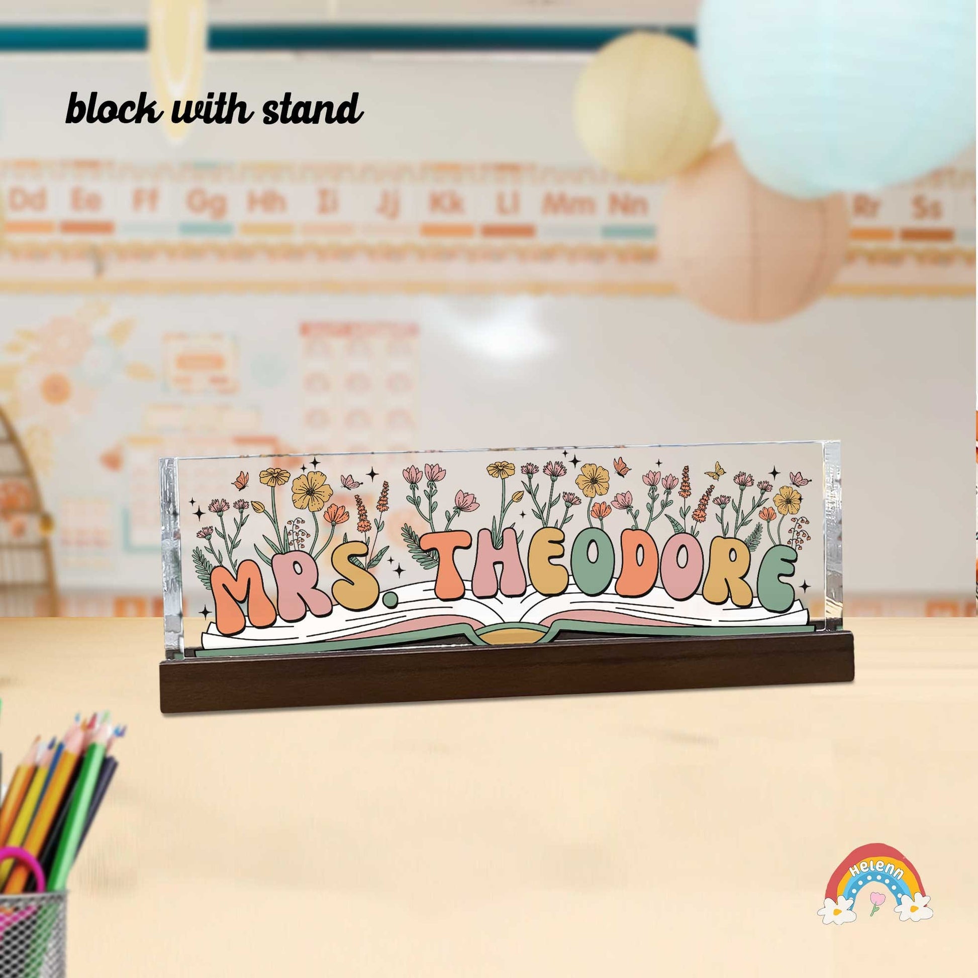 A decorative nameplate sits prominently on a desk, identifying Mrs. Theodore's workspace. It showcases a colorful, floral motif growing out of an open book, symbolizing a passion for teaching and nature.