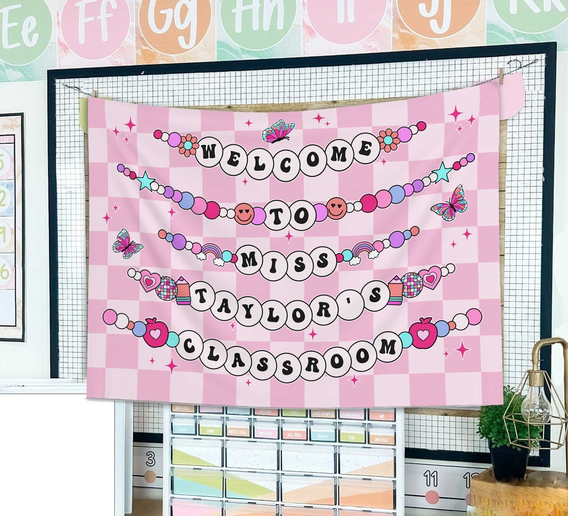 A welcoming banner with Welcome to Miss Taylor&#39;s Classroom in beaded letters on a pink checkered background with hearts, butterflies, and playful icons.
