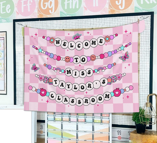 A welcoming banner with Welcome to Miss Taylor's Classroom in beaded letters on a pink checkered background with hearts, butterflies, and playful icons.