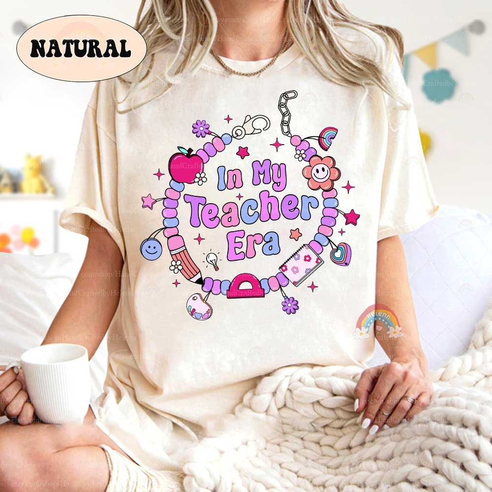This T-shirt showcases a playful theme with the words In My Teacher Era, highlighted by a beaded bracelet design adorned with fun and vibrant teacher-related charms. Perfect for celebrating a love of teaching.