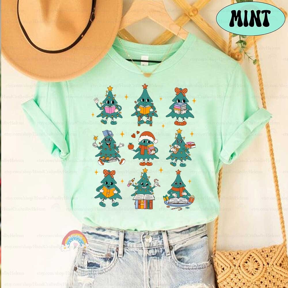 Adorable T-shirt featuring cute Christmas tree characters, each holding different school-related items like books and pencils. The festive design brings holiday cheer with stars and playful details.