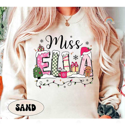 Charming holiday design showcasing Miss Ella’s name adorned with festive accents like Christmas lights, a Santa hat, and winter accessories. A joyful holiday sweatshirt for teachers.