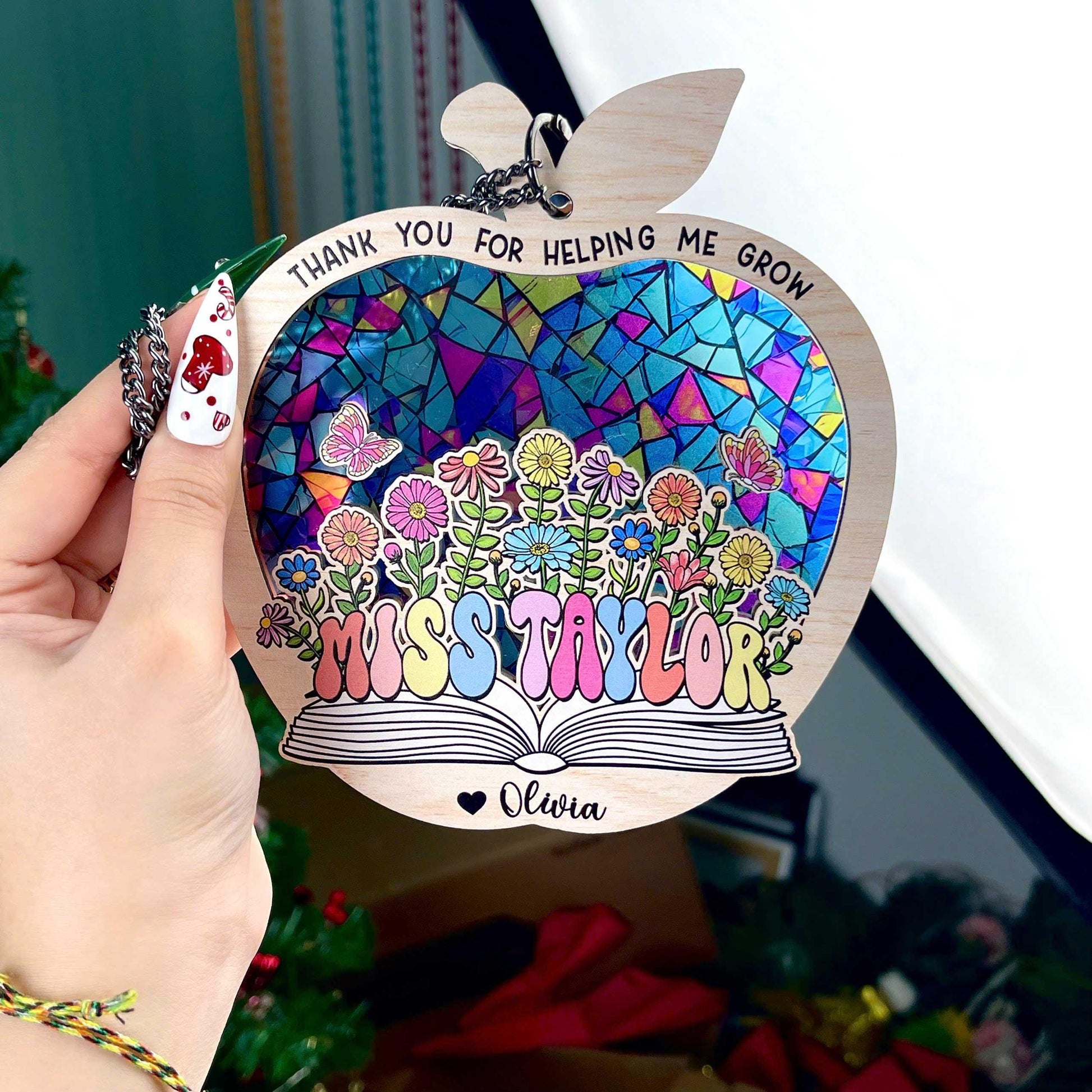 May include: Wooden apple-shaped teacher appreciation ornament.  Features a stained-glass style mosaic design in blues, purples, pinks, and greens.  Flowers and butterflies are depicted. Text includes: &#39;Thank you for helping me grow&#39;, &#39;Miss Taylor&#39;, and &#39;Olivia&#39;.  The ornament has a book shape at the bottom. Pastel rainbow-colored lettering is used for the teacher&#39;s name.