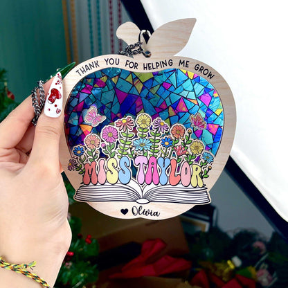 May include: Wooden apple-shaped teacher appreciation ornament.  Features a stained-glass style mosaic design in blues, purples, pinks, and greens.  Flowers and butterflies are depicted. Text includes: &#39;Thank you for helping me grow&#39;, &#39;Miss Taylor&#39;, and &#39;Olivia&#39;.  The ornament has a book shape at the bottom. Pastel rainbow-colored lettering is used for the teacher&#39;s name.