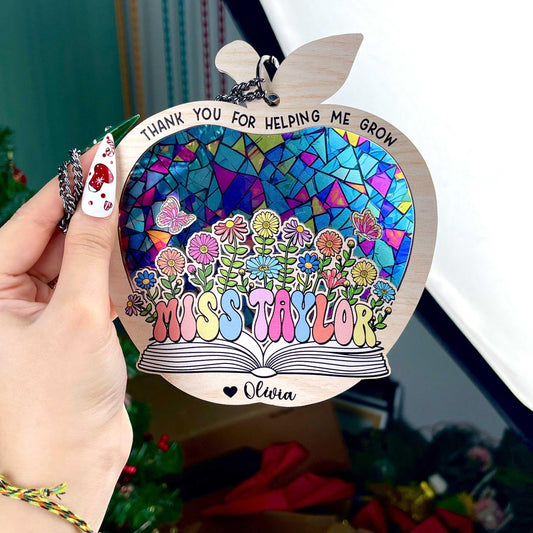 May include: Wooden apple-shaped teacher appreciation ornament.  Features a stained-glass style mosaic design in blues, purples, pinks, and greens.  Flowers and butterflies are depicted. Text includes: &#39;Thank you for helping me grow&#39;, &#39;Miss Taylor&#39;, and &#39;Olivia&#39;.  The ornament has a book shape at the bottom. Pastel rainbow-colored lettering is used for the teacher&#39;s name.