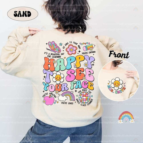 May include: A beige sweatshirt with a colorful graphic print on the back. The graphic includes the words 'Happy to see your face', a rainbow, a flower, a globe, an apple, and a butterfly. The front of the sweatshirt has a smaller graphic of a flower with a smiley face and the words 'Mrs. Smith'.
