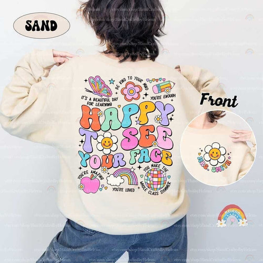 May include: A beige sweatshirt with a colorful graphic print on the back. The graphic includes the words &#39;Happy to see your face&#39;, a rainbow, a flower, a globe, an apple, and a butterfly. The front of the sweatshirt has a smaller graphic of a flower with a smiley face and the words &#39;Mrs. Smith&#39;.